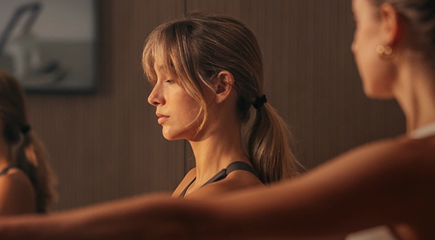 A woman in a side profile with a focused, calm expression during a PILAT3S class. She has blonde hair tied back and is wearing athletic wear.
